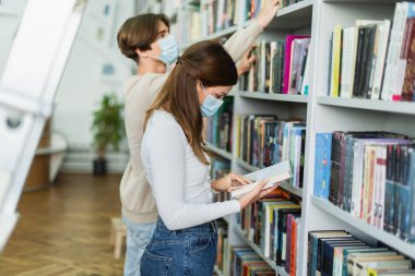 side view of teenage girl in medical mask reading book near rack and guy on blurred background