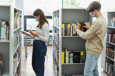 couple of teenage students in medical masks choosing books on bookshelves in library