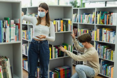 teenage girl in medical mask standing with cellphone near friend choosing books in library
