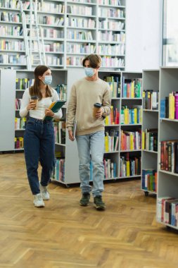 teenage students in medical masks walking with notebooks and laptop in library