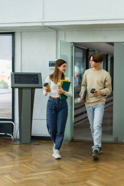 full length of teenage friends with coffee to go and notebooks entering library