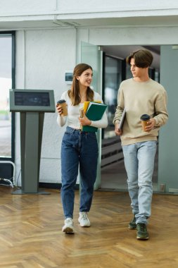smiling teenage girl with notebooks talking to friend with laptop while entering library