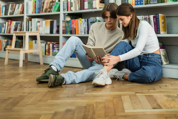 full length of teenage students sitting on floor near library racks and reading book