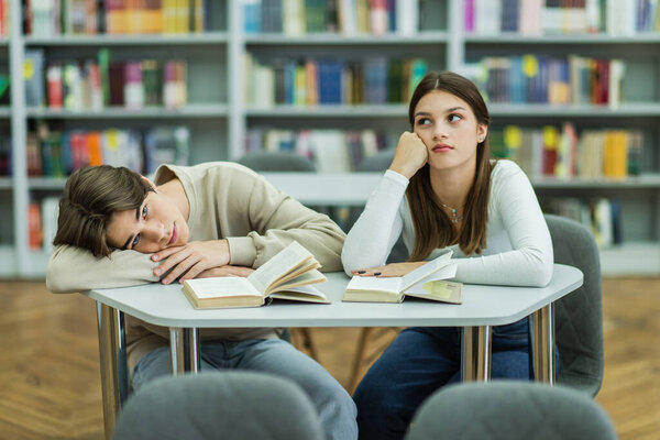 bored guy looking at camera near books and thoughtful teenage girl in library reading room