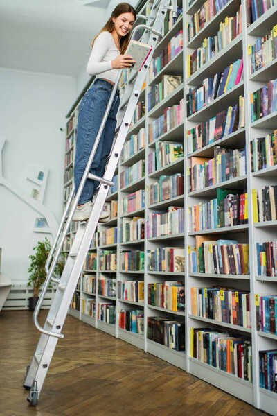 happy teenage girl holding book while standing on ladder in library