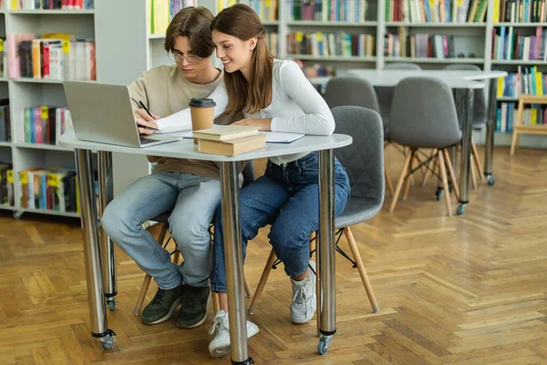 smiling teenage girl using laptop while studying with friend in library