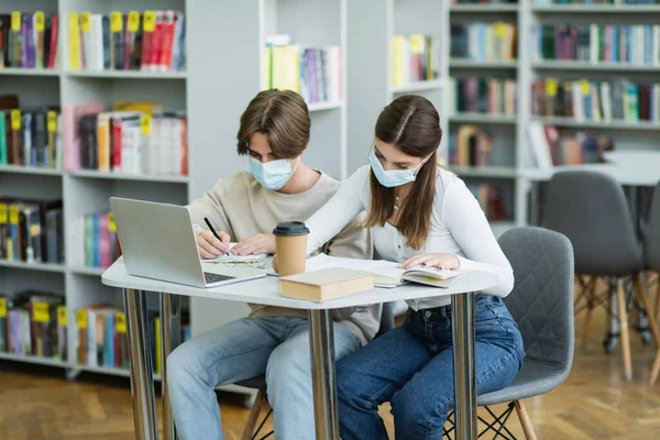 teenage students in medical masks studying near laptop and bookshelves in library