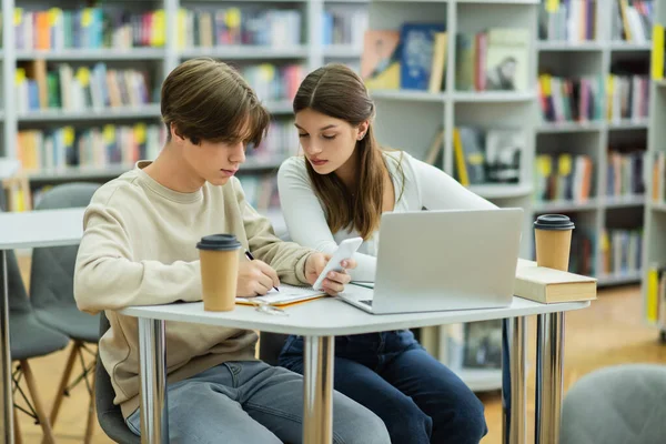 student holding smartphone and writing near laptop and teenage girl in library reading room