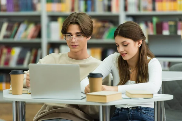 smiling teenage friends studying near laptop and coffee to go in reading room