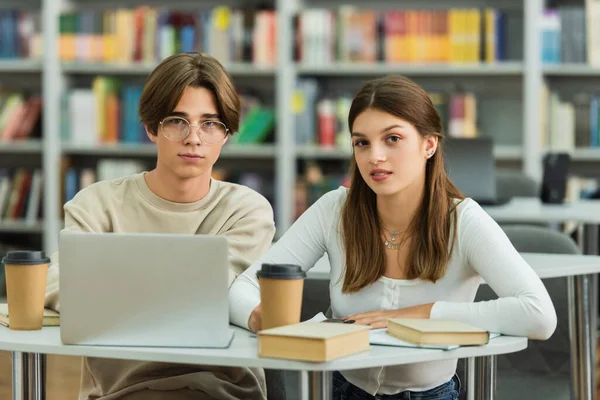 teenage friends looking at camera near laptop and coffee to go in library on blurred background