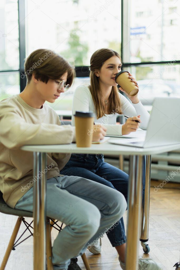 teenage girl drinking coffee near laptop and friend writing in library reading room