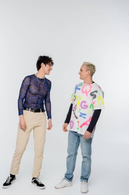trendy gay man and nonbinary person smiling at each other on grey background