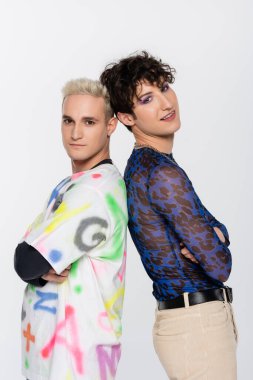 blonde gay man and smiling queer person standing back to back with crossed arms isolated on grey