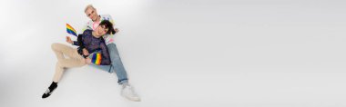 top view of happy nonbinary person and gay sitting with lgbtq flags on grey background, banner
