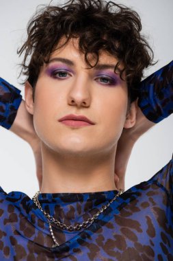 portrait of brunette queer person with makeup and hands behind head isolated on grey