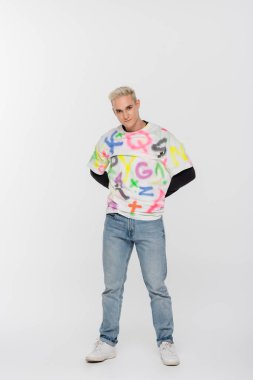 full length of gay man in jeans and t-shirt with alphabet print posing with hands behind back on grey background