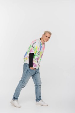 blonde gay man with hand in pocket of jeans looking at camera on grey background