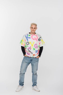 full length of trendy gay man standing with hands in pockets of jeans on grey background