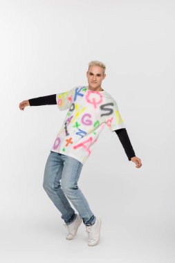 full length of gay in alphabet print t-shirt and jeans posing on tiptoes on grey background