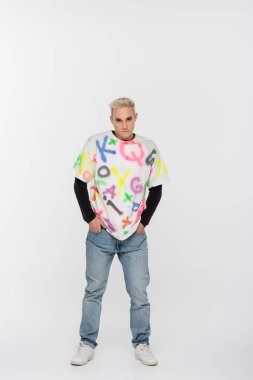 young gay man in alphabet print t-shirt standing with hands in pockets of jeans on grey background