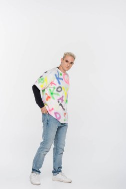 young and stylish gay man posing with hand in back pocket on grey background