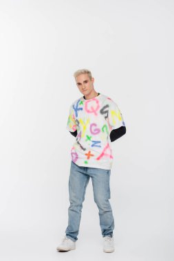 full length of young gay man in jeans and t-shirt with multicolored alphabet print on grey background