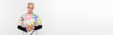 young blonde gay in stylish multicolored t-shirt looking at camera isolated on grey, banner