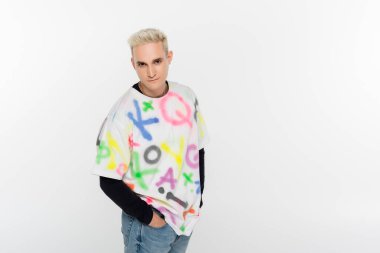 young gay man standing with hands in pockets and looking at camera isolated on grey