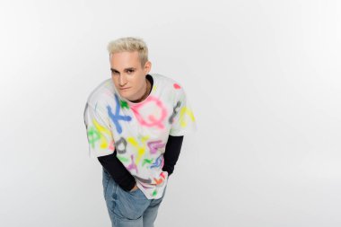 blonde gay man looking at camera while posing with hands in pockets of jeans isolated on grey