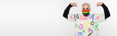 blonde gay in alphabet print t-shirt and lgbtq colors medical mask showing strength isolated on grey, banner