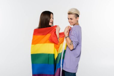 tattooed woman smiling near girlfriend wrapped in lgbtq flag isolated on grey