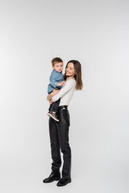 full length of young and happy woman holding toddler son on grey background