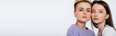 blonde woman with makeup looking at camera near young brunette girlfriend isolated on grey, banner