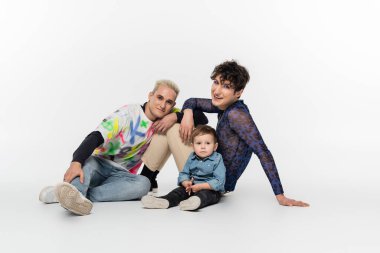 happy gay couple smiling at camera near toddler son on grey background
