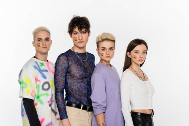 young and stylish people of different identity and with rainbow flags on faces looking at camera isolated on grey
