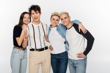 happy man showing victory sign near trendy friends hugging and looking at camera isolated on grey