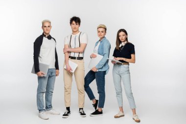 full length of young and fashionable friends with laptops and digital tablets on grey background