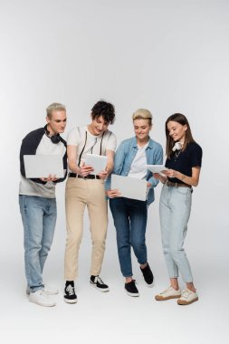 full length of stylish friends using laptops and digital tablets on grey background