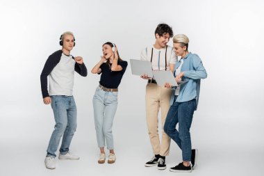smiling couple using laptops near cheerful friends dancing in wireless headphones on grey background