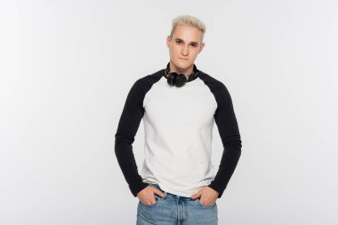 blonde man with headphones holding hands in pockets of jeans isolated on grey
