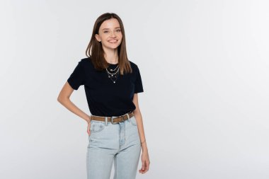 happy woman in black t-shirt and jeans posing with hand on hip isolated on grey