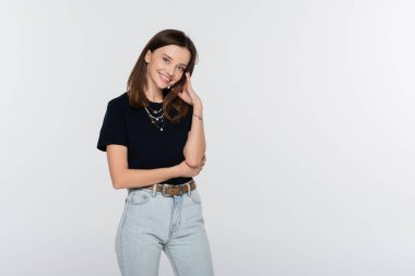 stylish brunette woman standing with hand near face and smiling at camera isolated on grey