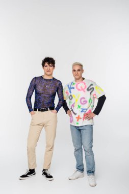 full length of positive gay man and queer person in stylish clothes posing on grey background