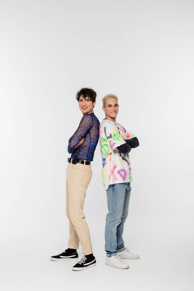 full length of cheerful gay man and queer person standing back to back with crossed arms on grey background