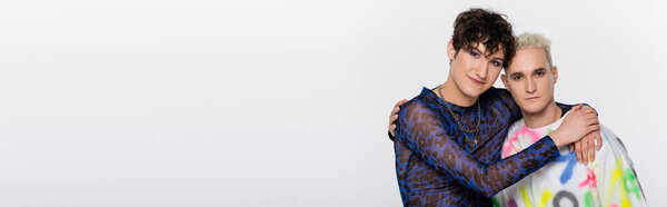 brunette nonbinary person and blonde gay man hugging and looking at camera isolated on grey, banner