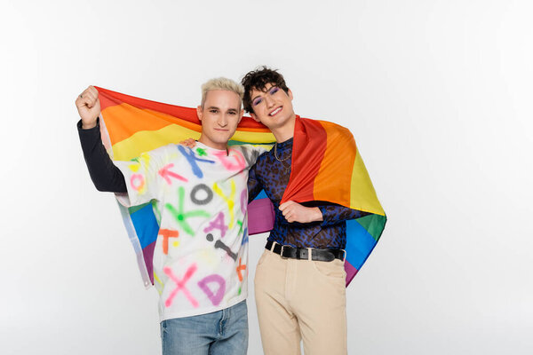 young and stylish gay man and queer person with lgbt flag smiling at camera isolated on grey