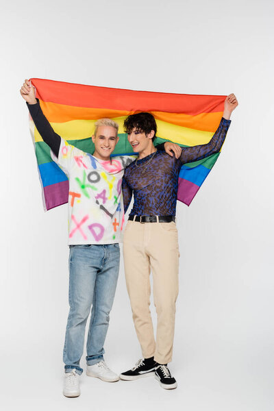 full length of gay man and nonbinary person holding lgbtq flag on grey background