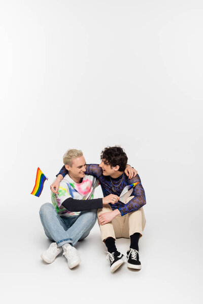 happy gay man and nonbinary person with small lgbtq flags embracing while sitting on grey background
