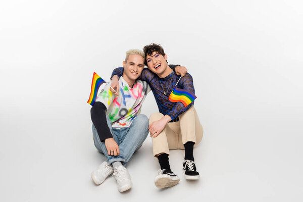 high angle view of smiling gay man and queer person sitting with lgbtq flags on grey background
