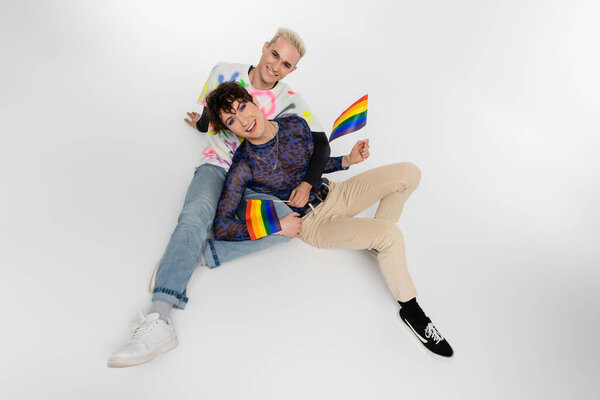 joyful nonbinary person with small lgbtq flags sitting near smiling gay man on grey background